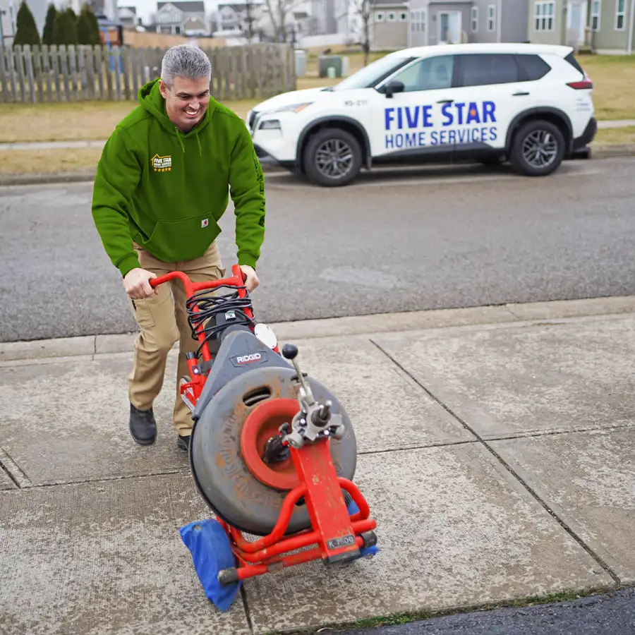 Plumbing wheeling plumbing equipment up a driveway with a five star home services truck behind him.